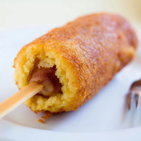 Golden brown corn dogs resting on a paper towel-lined plate, with a bowl of ketchup and yellow mustard for dipping.